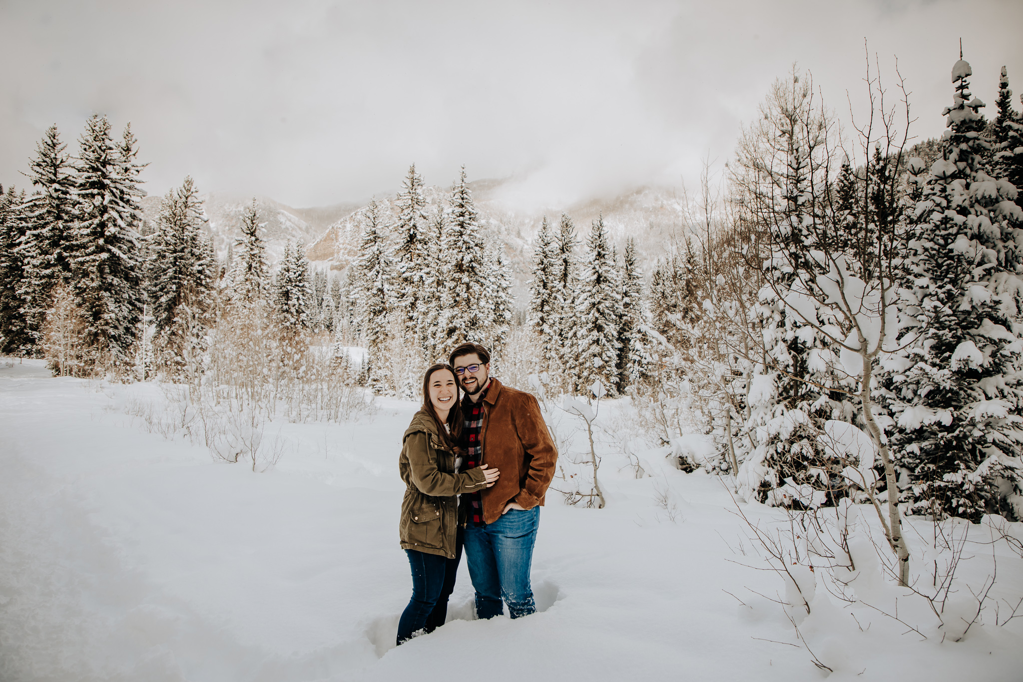 A couple facing the camera and embracing. They are surrounded by snow covered trees and mountains. They're both smiling at the camera