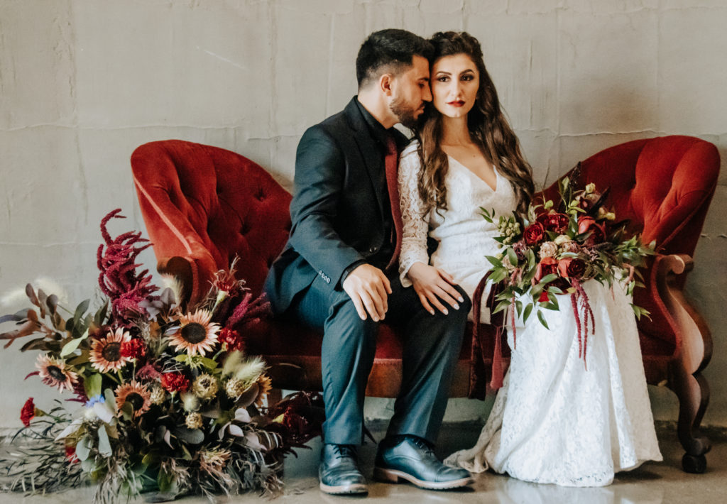 A married couple sitting on burgundy couch with two large floral arrangements, the bride is looking at the camera while the groom nuzzles her cheek