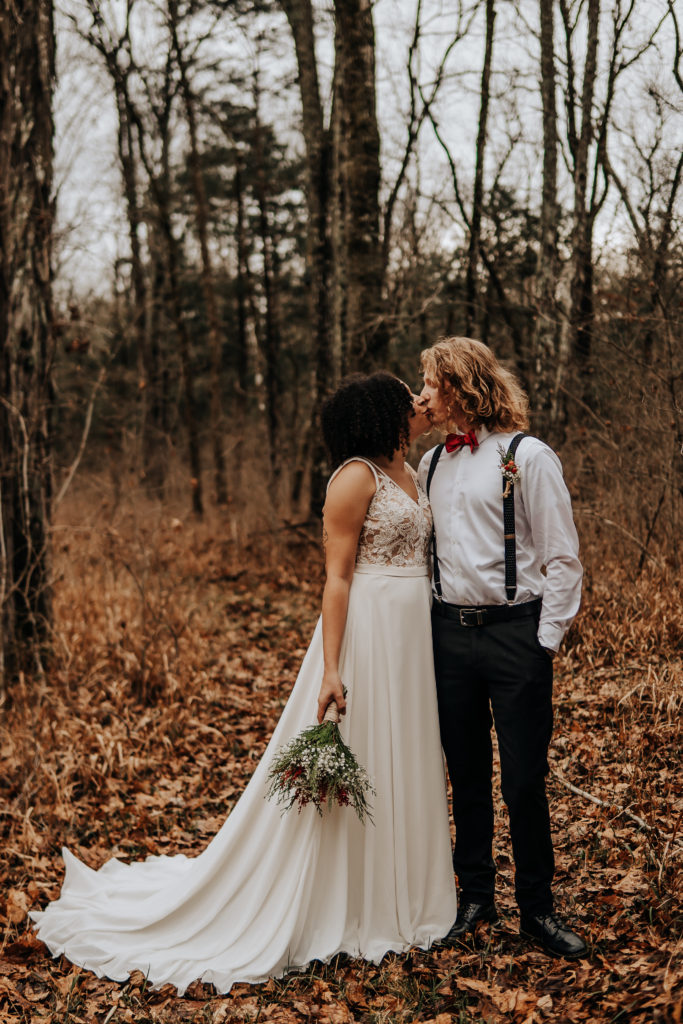 A couple standing the woods in their wedding attire, they are facing each other and kissing. Her entire dress is laid out perfectly against the backdrop