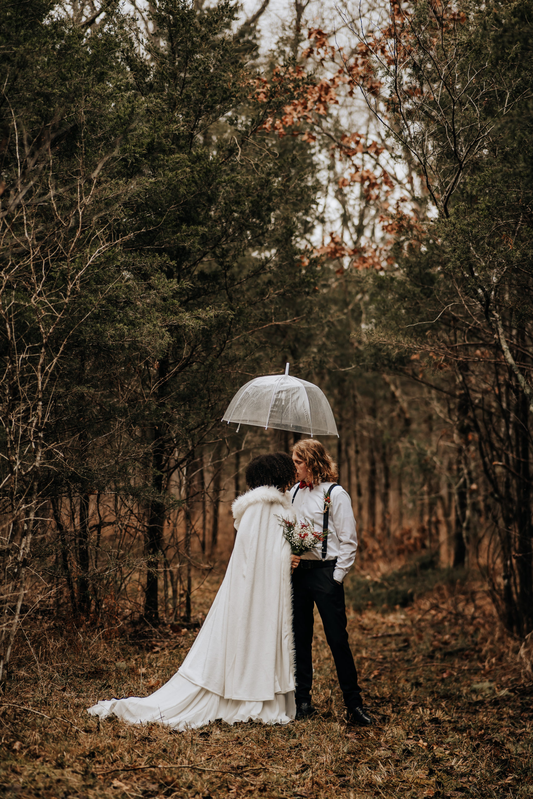 A couple standing facing each other, they are wearing their wedding attire, the bride has a beautiful white cape on with a hood. You can tell it's raining because they are holding a clear umbrella above their heads. You can see their entire bodies as well as the wooded landscape that surrounds them