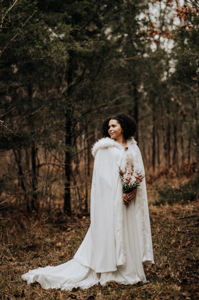A woman in her wedding attire, she has a beautiful white cape on and is holding her flowers and looking off into the distance. The frame captures her entire body and dress as well as the wooden landscape around her.