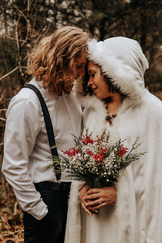 A close up image of a couple touching their foreheads together. They are dressed in their wedding attire and the bride has a beautiful white cape with a hood over her head and she is holding red and white flowers. The groom has his right hand in his pocket and a red bow tie. Both of their eyes are closed and they have a soft smile on their faces