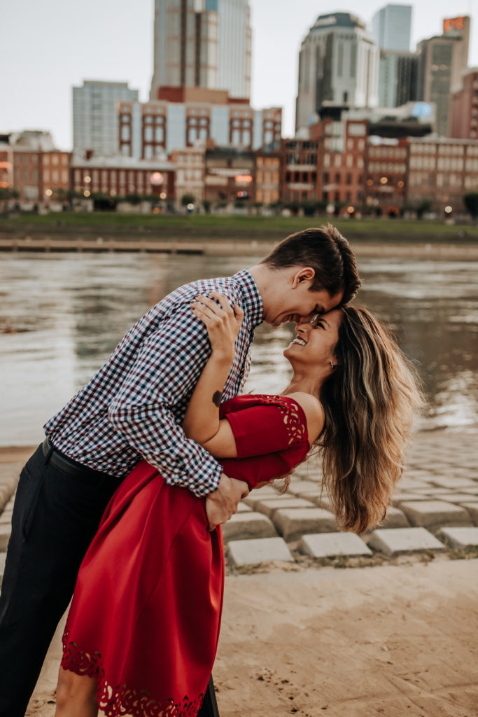 A straight couple holding each other. They are. touching foreheads and leaning back, they are about to kiss and there is a city skyline and a river behind them 