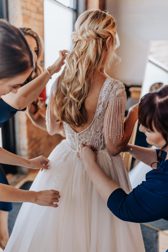 Minnesota wedding photo of a bride getting ready with her attendants. She is standing in front of a mirror wile her attendants fix her dress. The shot is from the back where we can see the brides hair and details of her dress. 