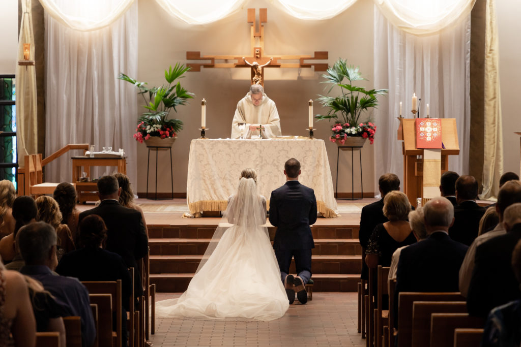 Bride & Groom kneeling at the foot of the alter while the priest prays over them at the top of the alter at Holy Name Catholic Church in Wayzata Minnesota