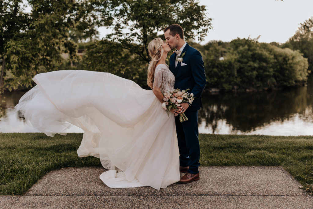 Minnesota wedding photograph of a couple kissing while the brides dress is in the air. They are standing in a park with a river behind them. They are both holding the floral bouqet