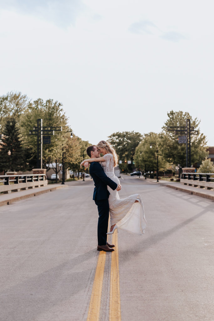Minnesota wedding photograph of a bride and groom standing in the middle of the road. The groom has picked up the bride and their foreheads are touching. The bride has a foot kicked out and they are smiling at each other. 