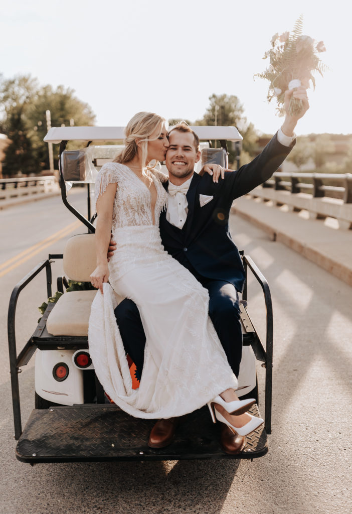 Bride & Groom taking their wedding portraits in Minnesota. They are sitting on the back of a golf cart with the bride in the grooms lap while she is kissing his cheek. The groom is looking at the camera and smiling while holding the bouquet in the air