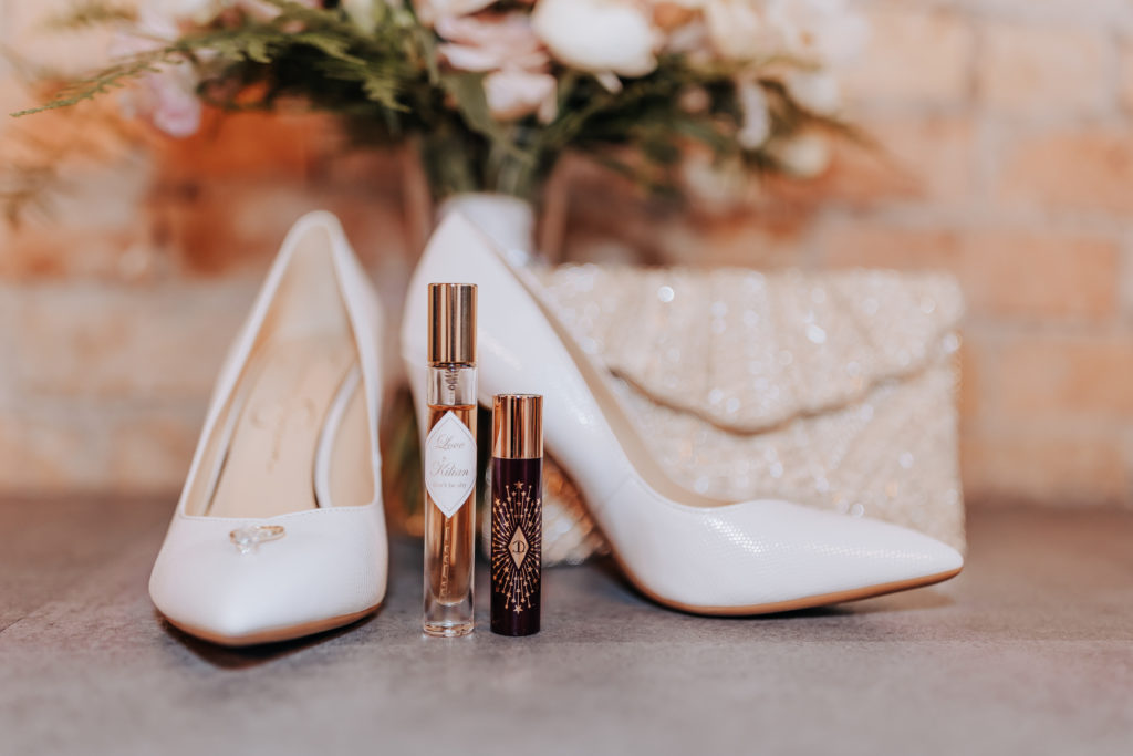 White stiletto heels with two small perfume bottles in front of them and a bouquet of flowers and small sequined bag rested against a brick wall and concrete floor at The Jerome Event Center in Delano, MN