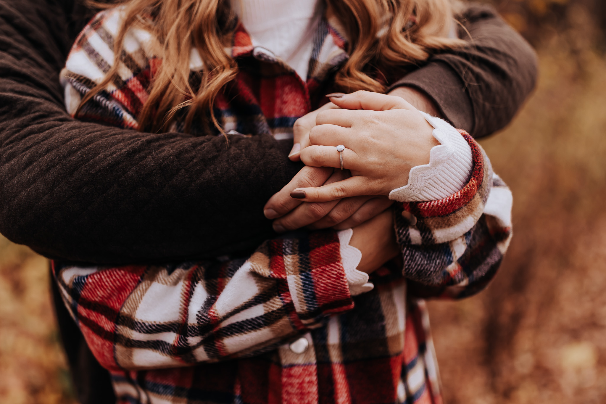 Detailed shot of the bride-to-be’s hand with a solitaire ring, framed by warm plaid flannel and layered sleeves.