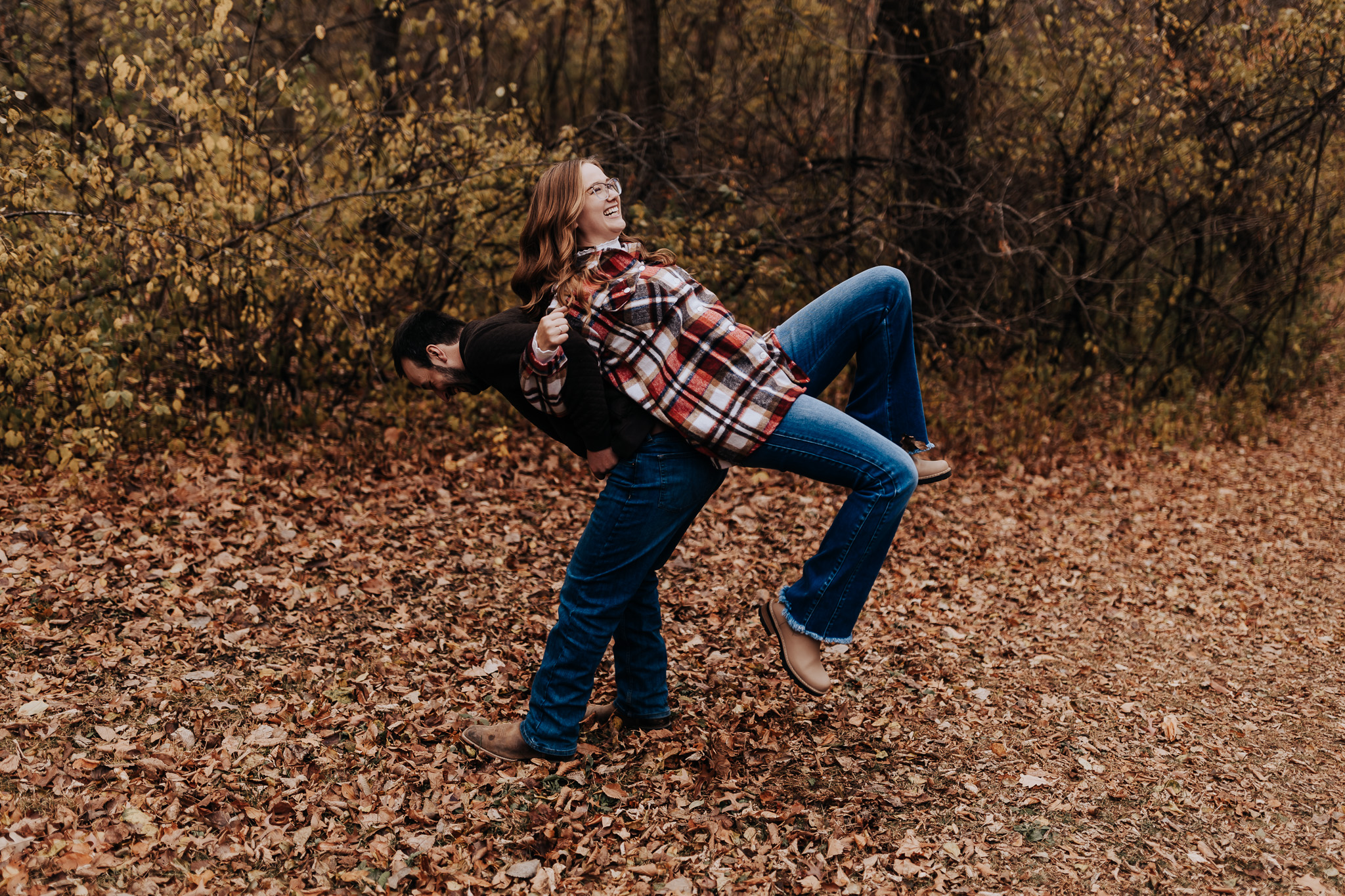 With leaves crunching underfoot, the couple laughs as he lifts her in a fun, unposed moment.