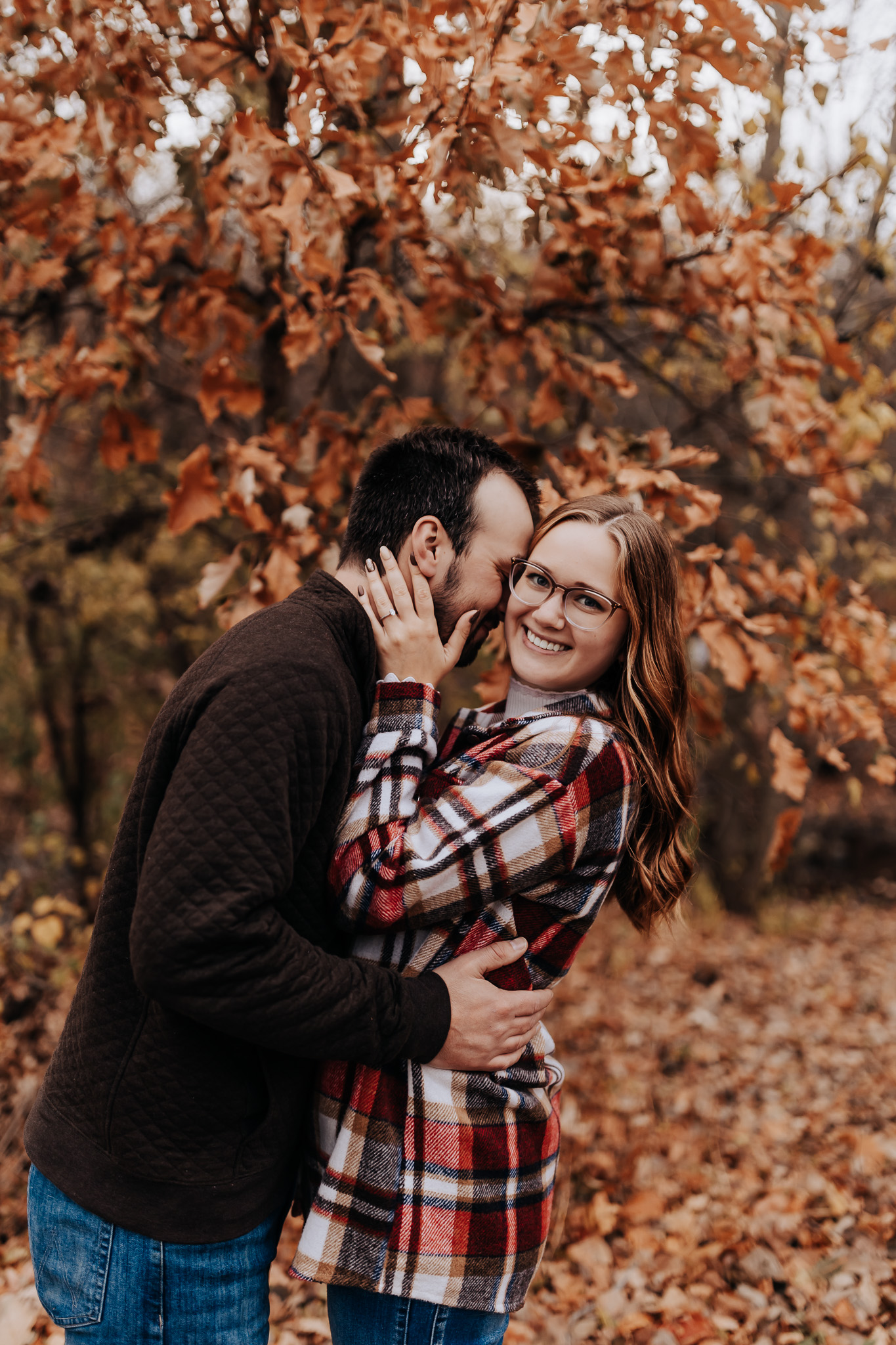 A straight couple's engagement photos. The male is holding his fiance and leaning into the side of her face. She is holding his neck in her hands and smiling at the camera. Minnesota Engagement Photographer.
