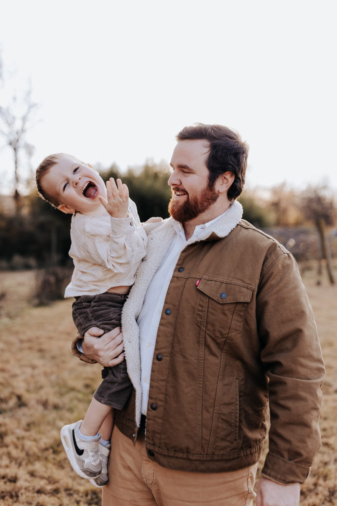 A young boy and his father. He's laughing at the camera while his father is carrying him and smiling at the boy. Nashville Family Photographer