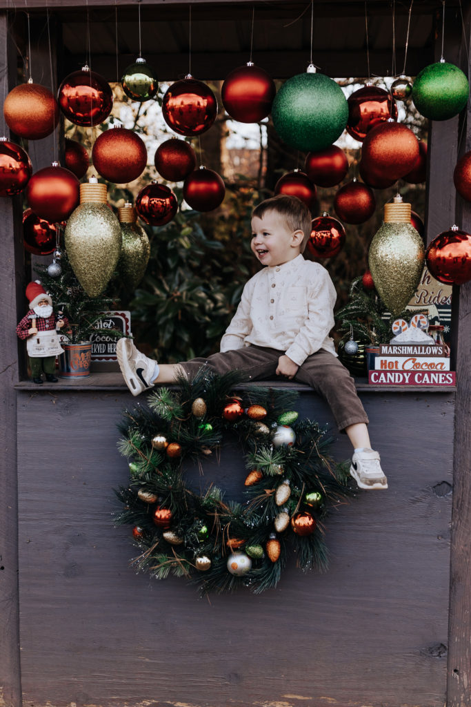 A young boy looking away from the camera. He is sitting with one leg dangling off a wooden platform surrounded by Christmas ornaments and decor. He is smiling. Nashville Family Photographer
