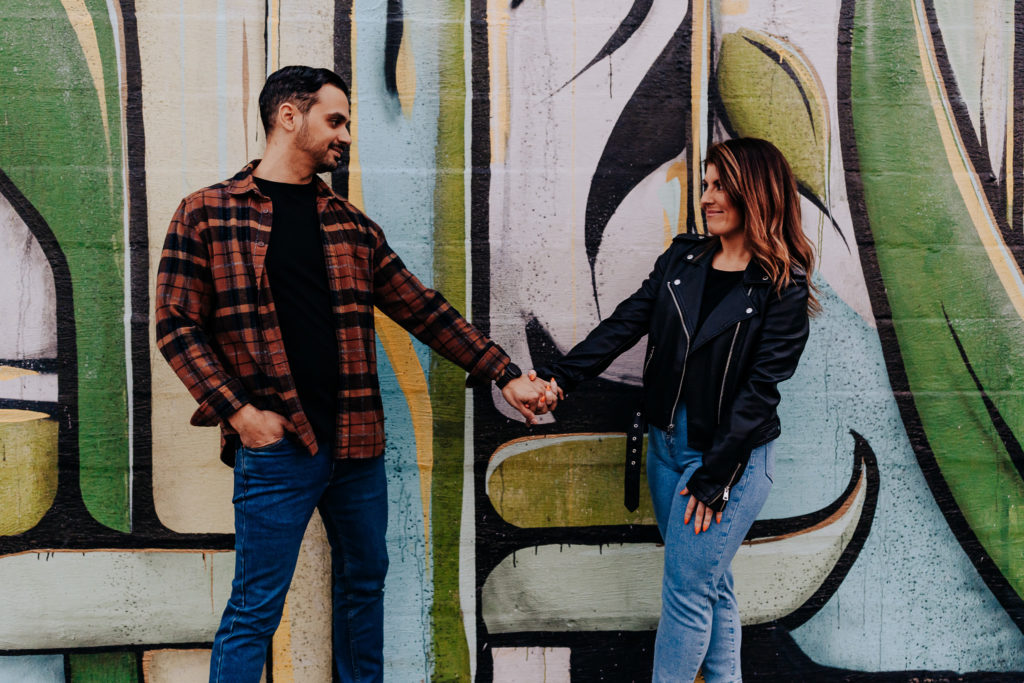 An engagement photo of a couple holding hands and standing a few feet away from each other. They are looking at each other and smiling. There is a green abstract mural behind them. Nashville rooftop engagement photographer