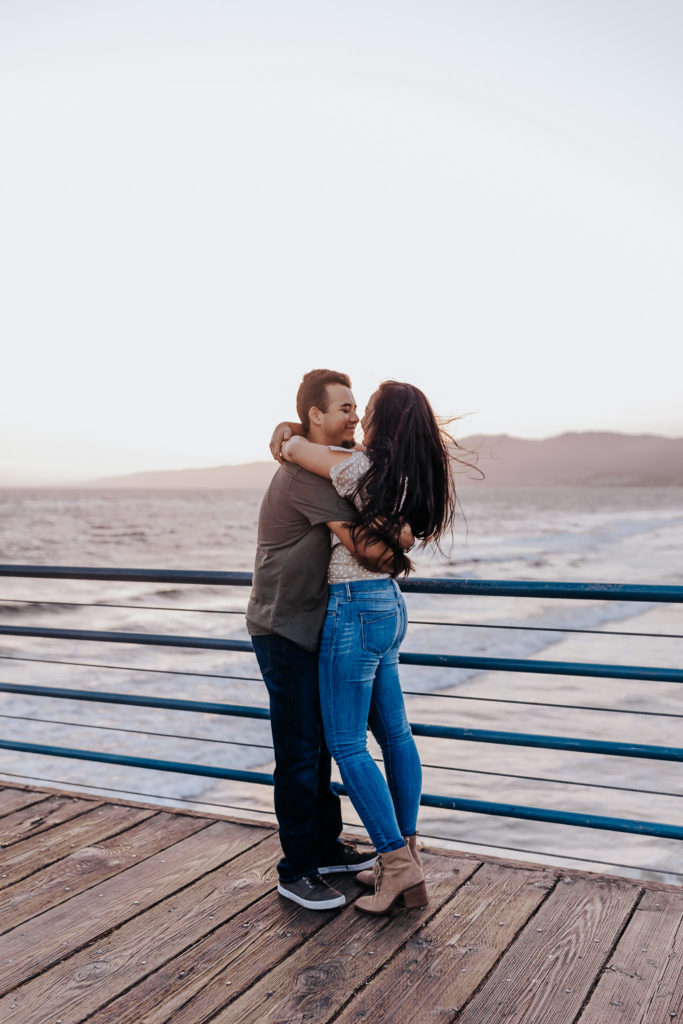 A straight couple holding each other. They are looking at each other as the girls hair blows in the wind. There is a sunset and ocean behind them. Santa Monica Pier Engagement Session