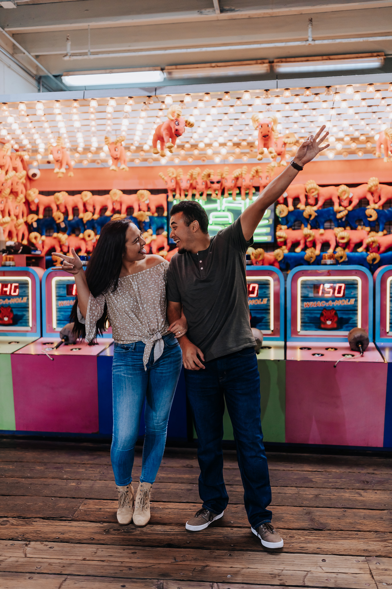 An engagement photo of a couple in front of a carnival game they are looking at each other and have their hands in the air. Both are smiling. Santa Monica Pier Engagement Session