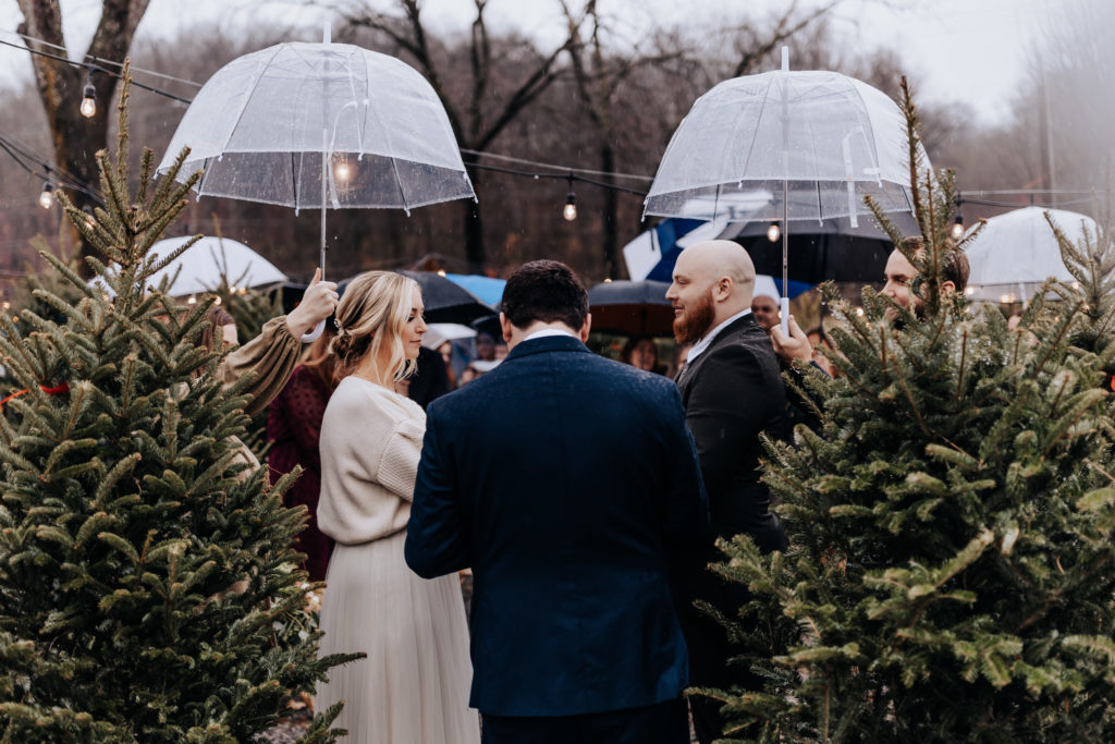 Couple about to get married. They are facing each other in front of their officiant. There are christmas trees around them. it is raining and two people are holding clear umbrellas over their heads. It is an intimate wedding at a christmas tree farm