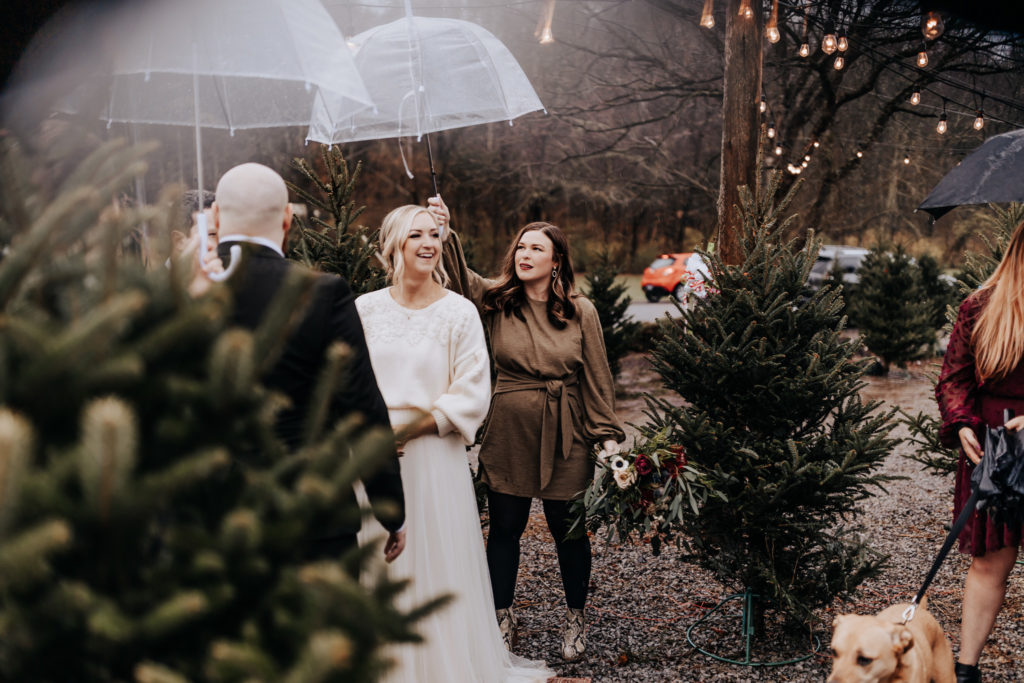 A couples standing in front of their friends and family. The best man and matron of honor are holding umbrellas over their heads because it's raining. They are smiling and about to get married. Christmas tree farm elopement.
