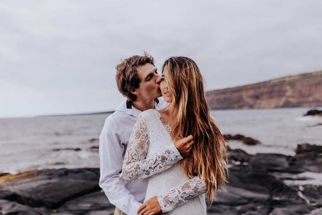 a groom kissing his bride on the cheek while she holds her hair. big island beach elopement