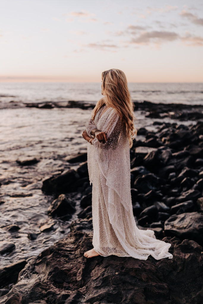 a bride is standing on the rocks by the ocean. she is looking off at the sunset and holding her arms. big island beach elopement