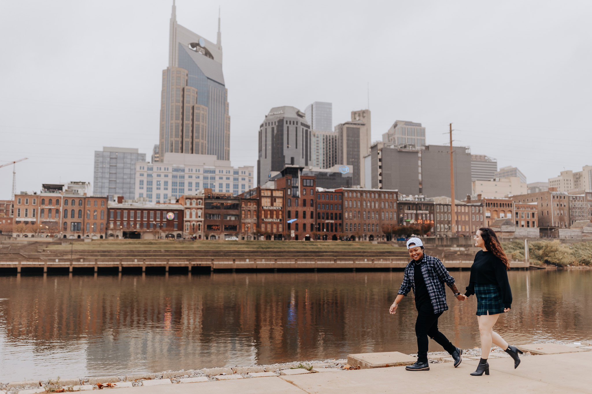A couple walks hand-in-hand along the Nashville waterfront, showing what to wear for engagement photos in the city with coordinated plaid and casual layers.