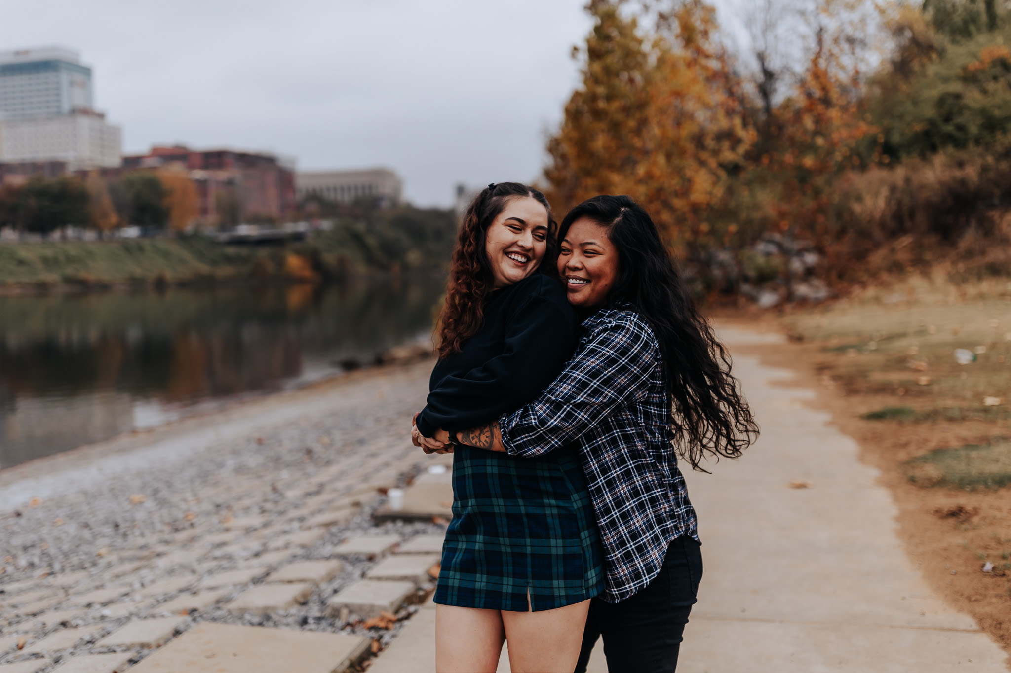Two women share a joyful hug on a riverside walkway during their fall engagement session.