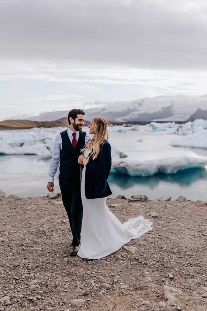 Iceland elopement photographer captures groom and bride walking beachside while bride wears groom's jacket