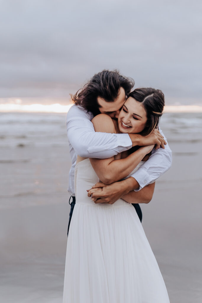 Destination wedding photographer captures groom hugging bride from behind on beach after Oregon Coast elopement