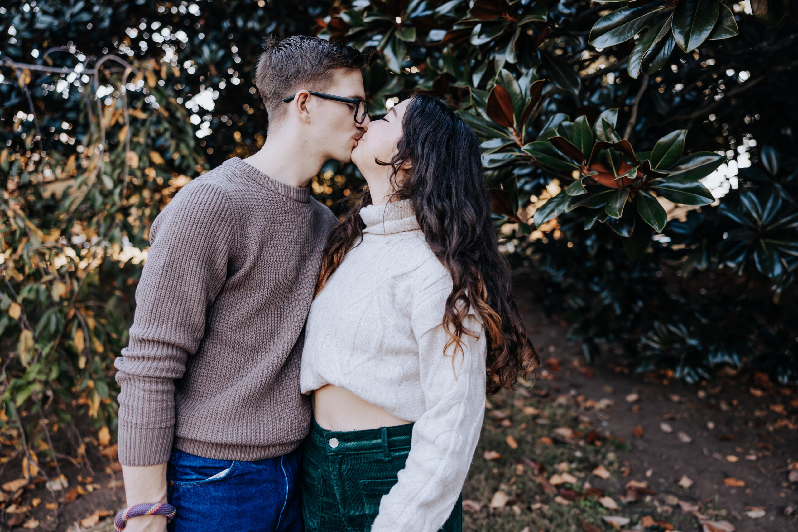 A couple kisses beneath magnolia trees, styled in cozy sweaters and corduroy for a warm fall engagement shoot.