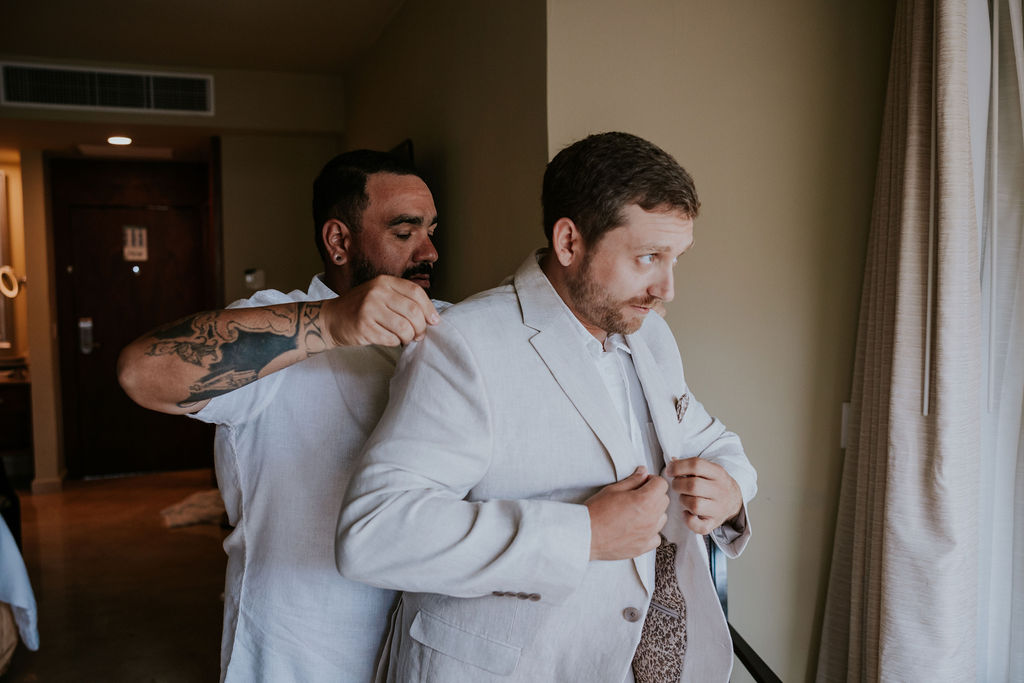 Destination wedding photographer captures groomsmen helping groom put on jacket