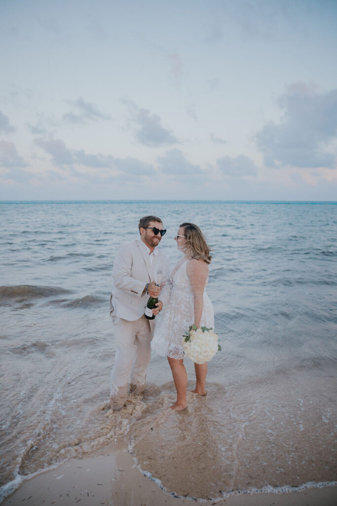 Destination wedding photographer captures bride and groom standing in ocean popping champagne