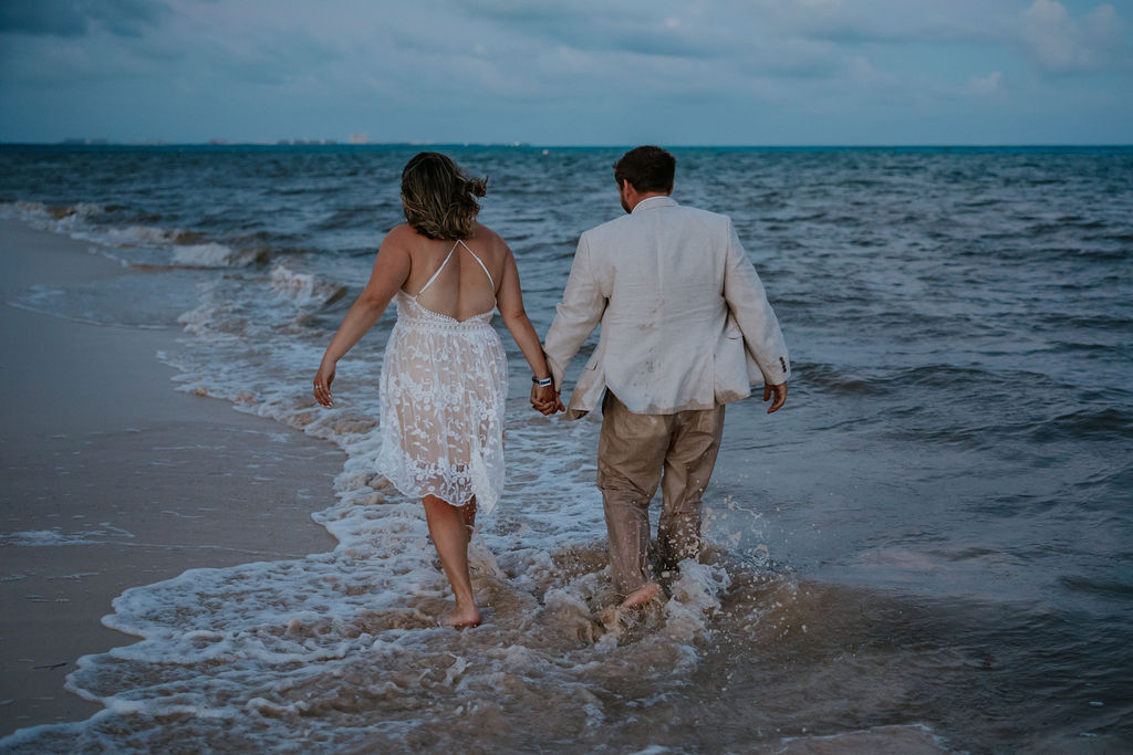 Destination wedding photographer captures bride and groom holding hands and walking through ocean