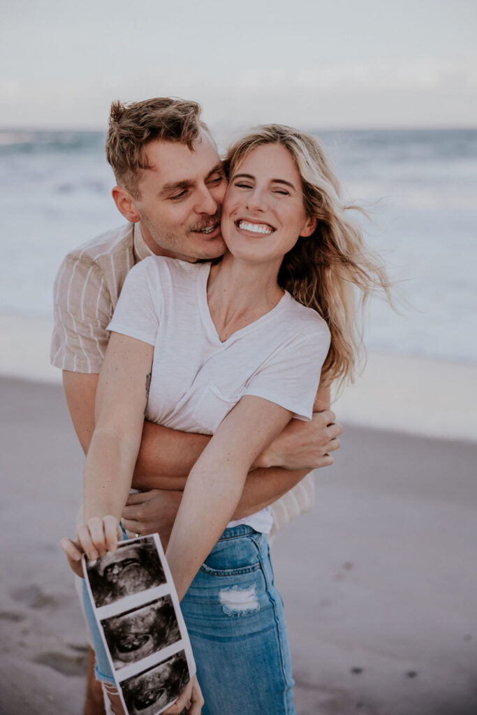 Destination Wedding Photographer captures man hugging woman from behind while she holds ultrasound