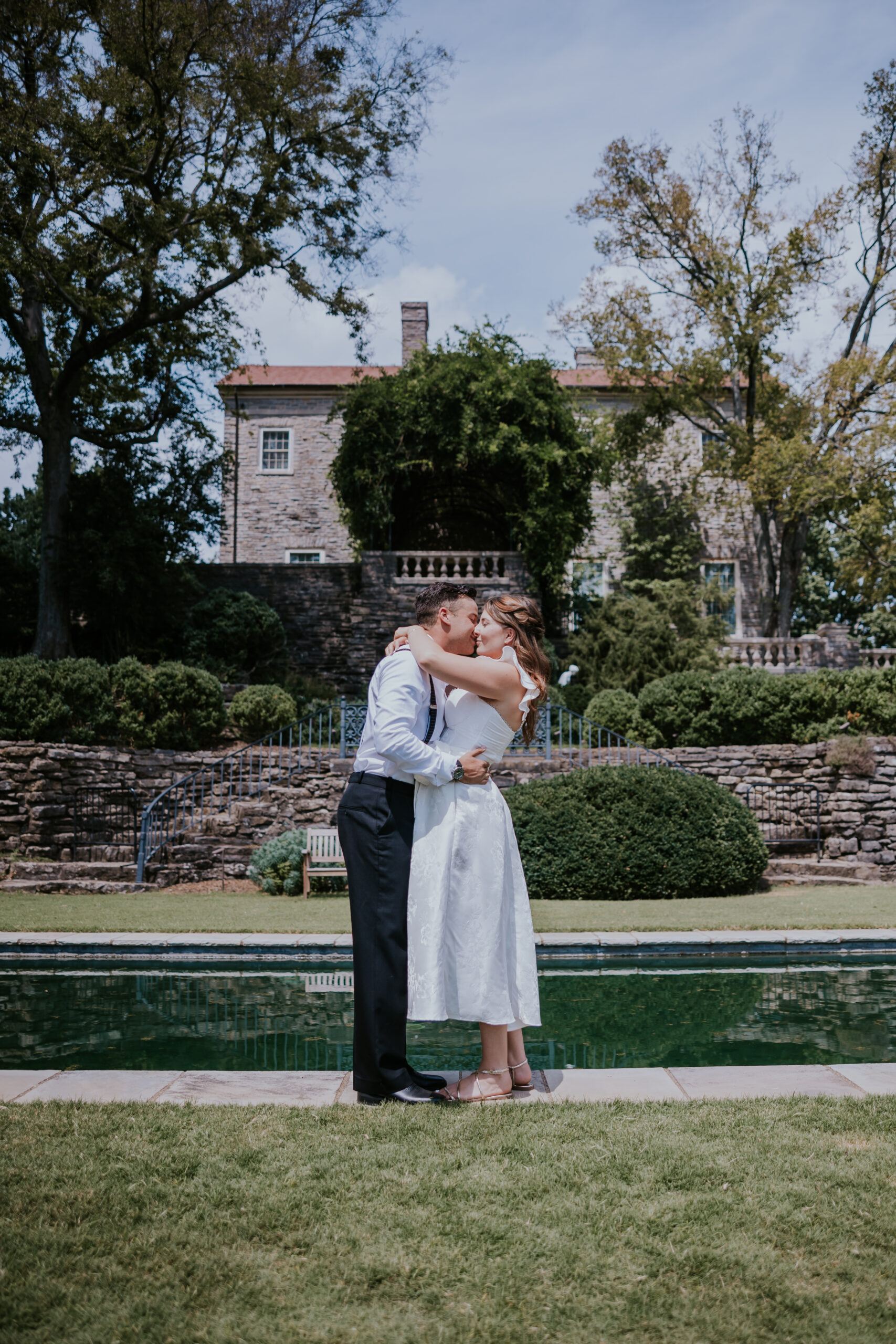 The couple shares a kiss in front of a historic stone estate, surrounded by gardens and a reflecting pool during their Nashville wedding.