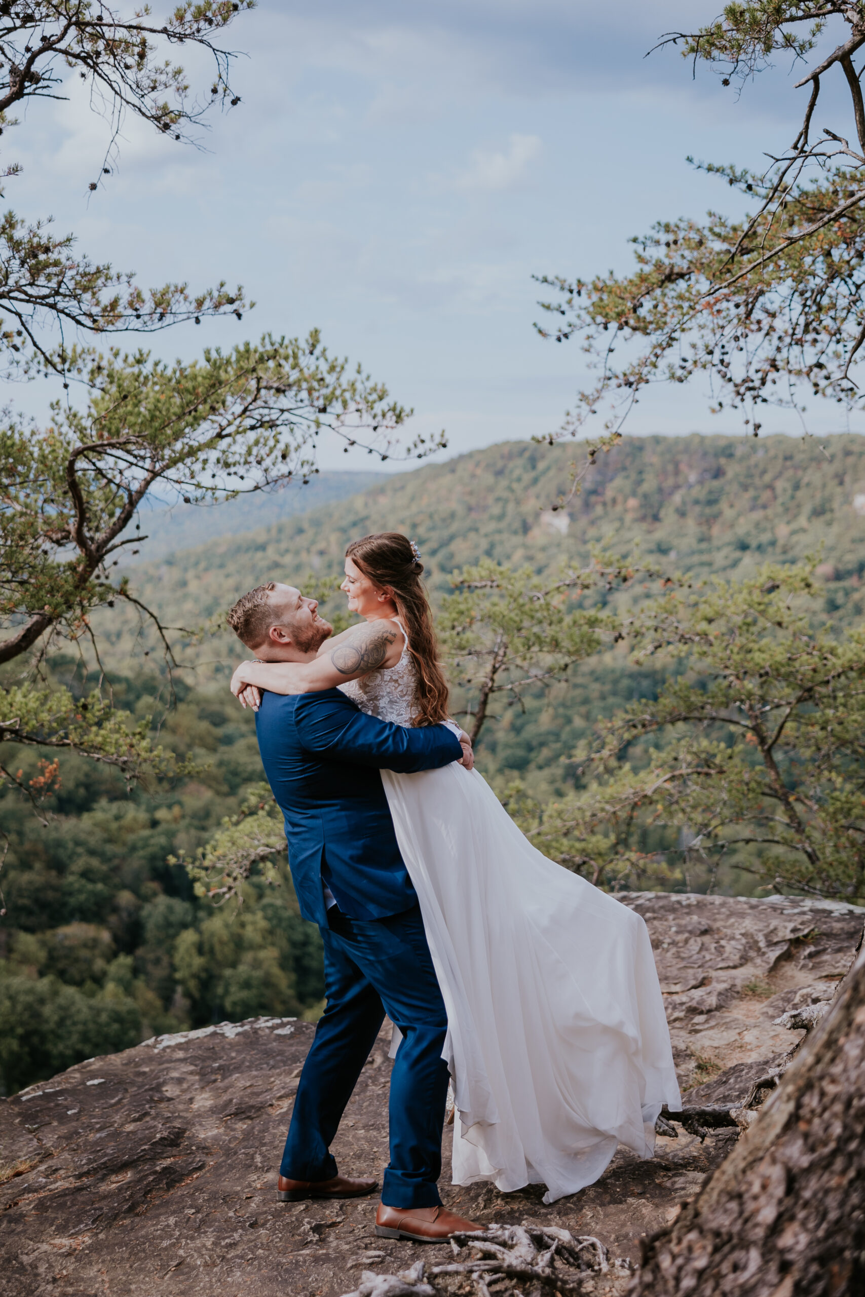 The groom lifts the bride on a scenic mountaintop ledge, surrounded by sweeping views and fall colors.
