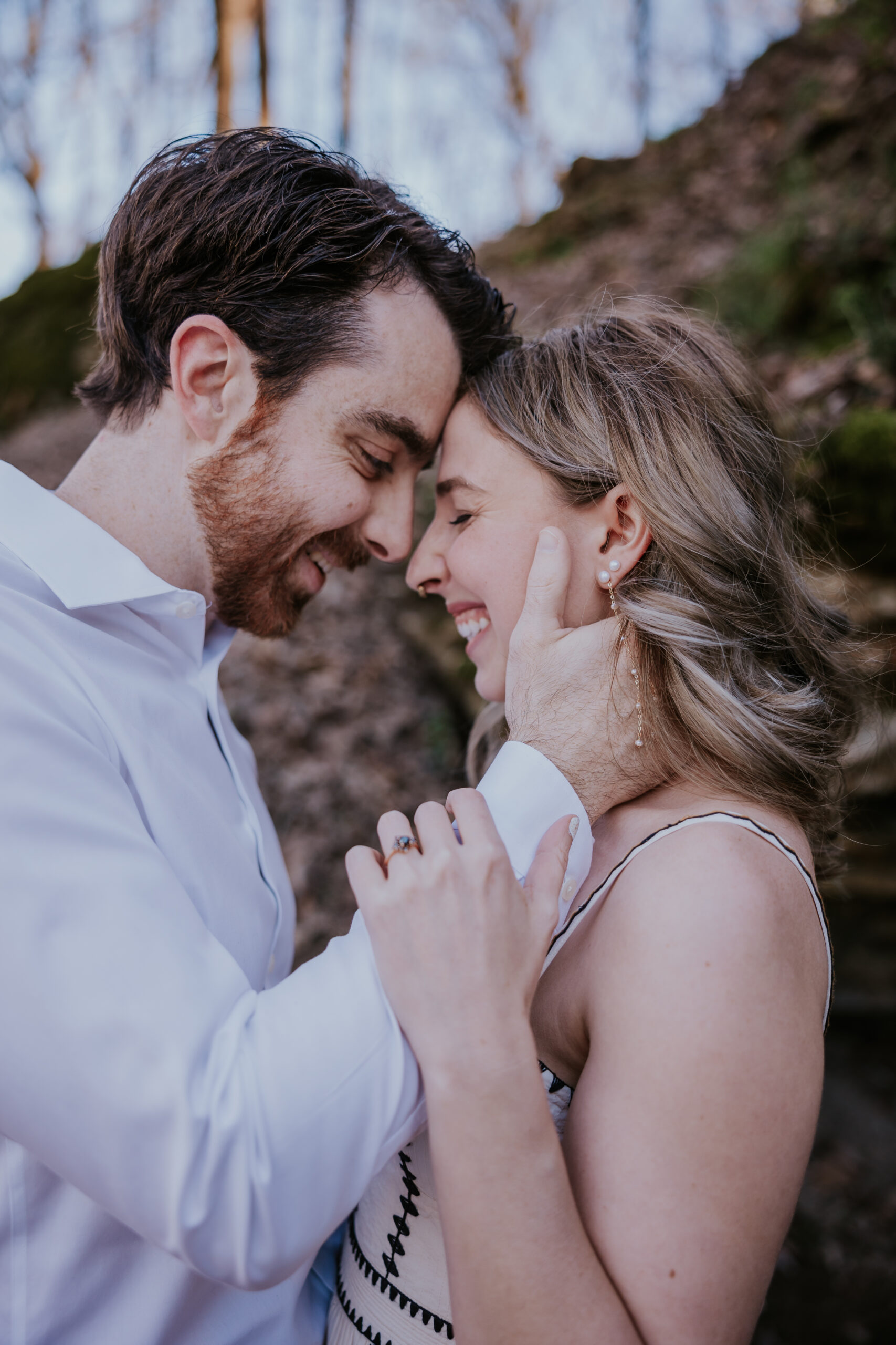 A couple shares a quiet, intimate moment in the woods, smiling with their foreheads pressed together.