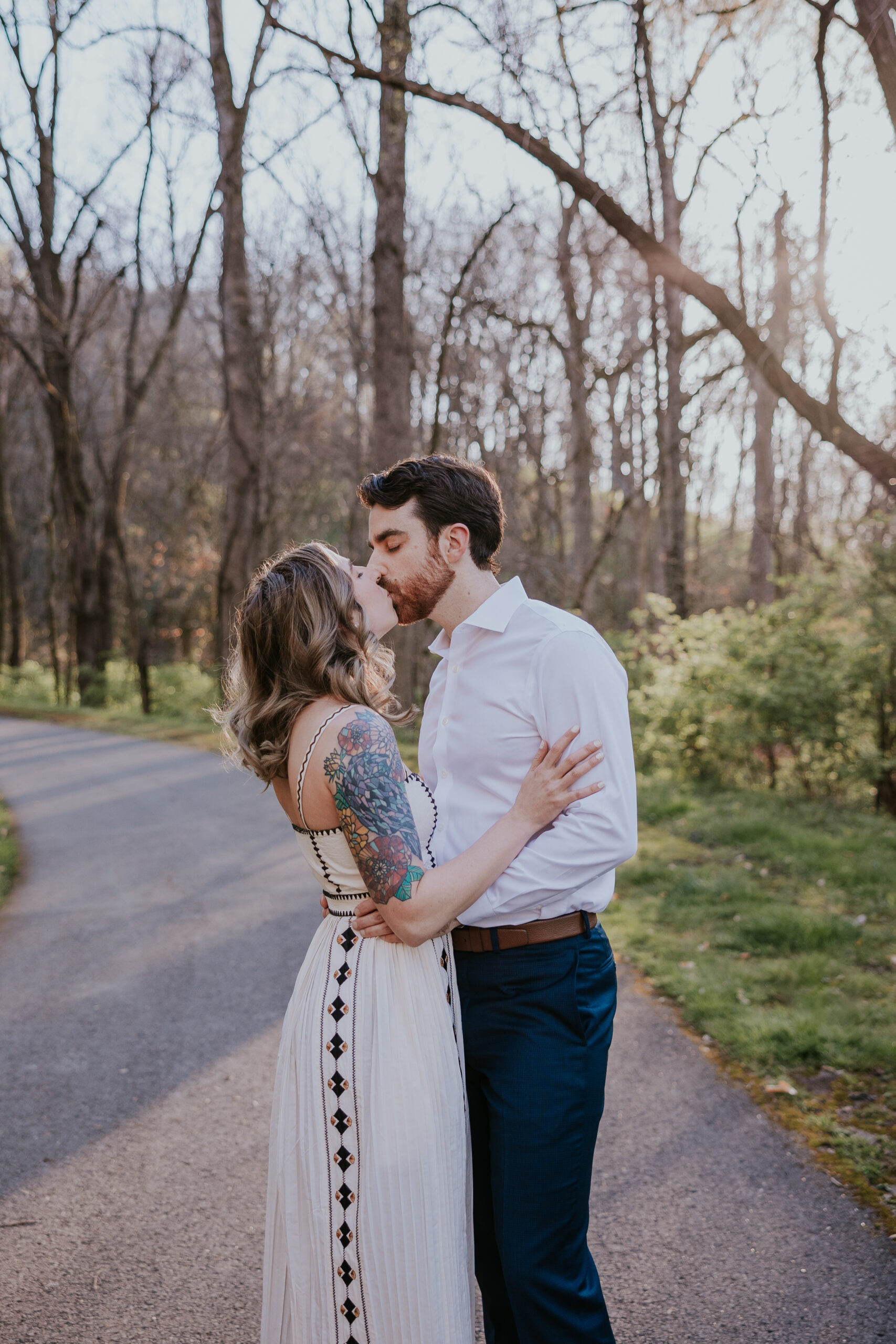 Bride with tattoos kisses groom on a forest path during golden hour, capturing a dreamy elopement vibe.