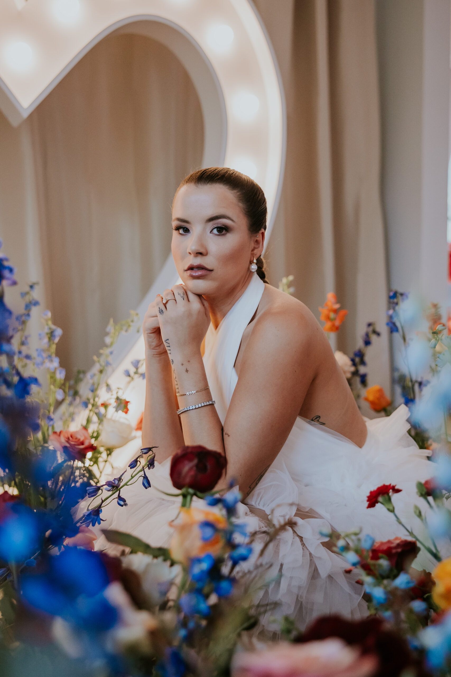 Elegant bride posing in a tulle gown surrounded by vibrant wildflowers and a heart-shaped marquee light, highlighting romantic wedding styling.
