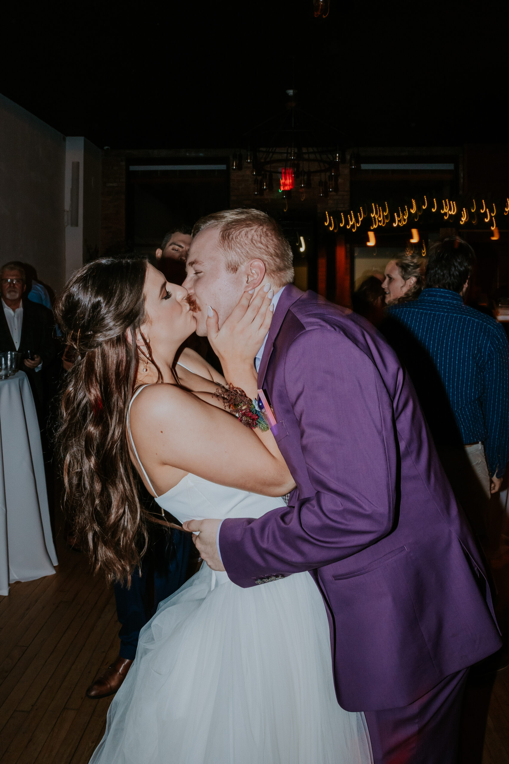 Bride and groom share a joyful kiss during their wedding reception, surrounded by guests and twinkle lights.