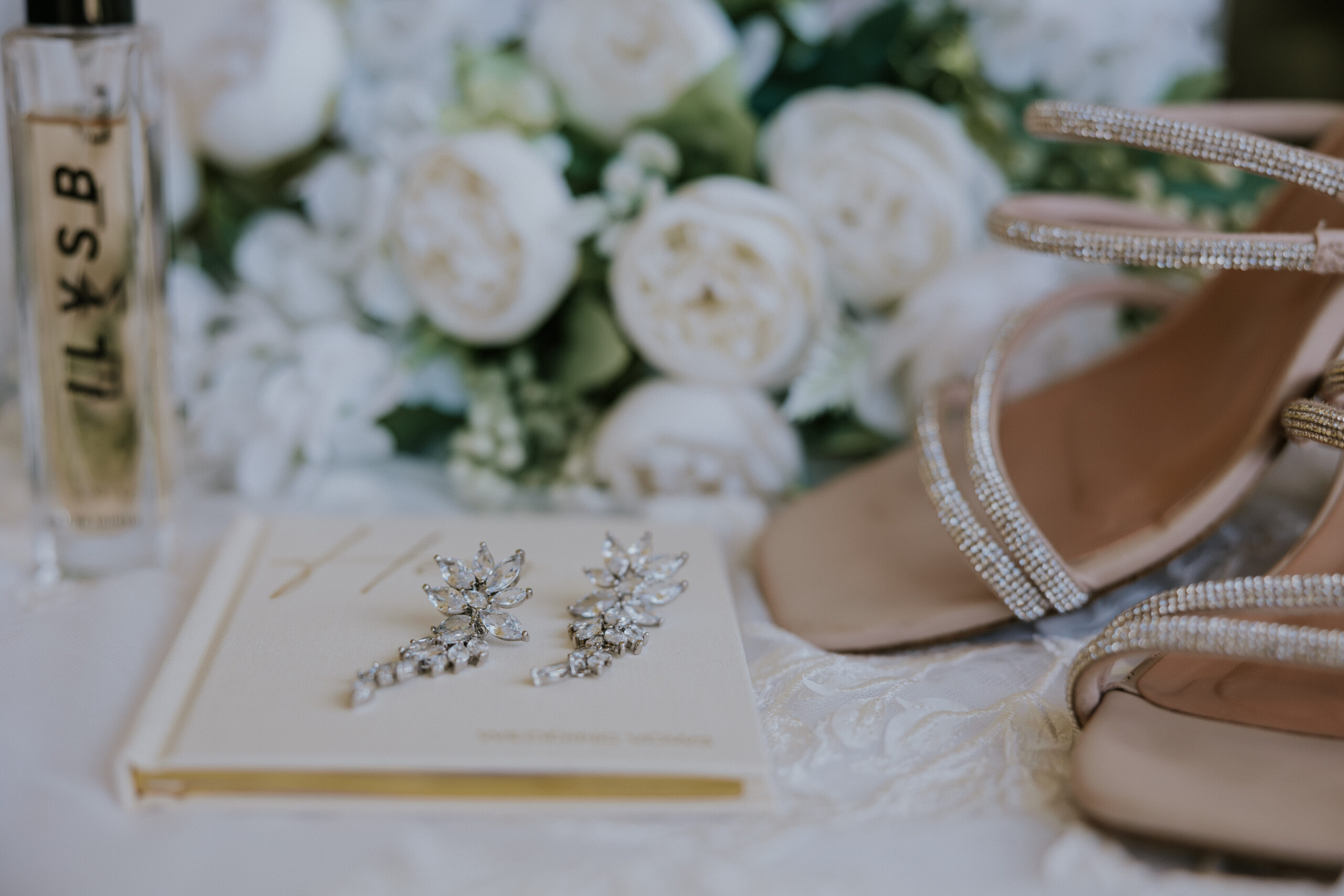 Bride’s sparkly heels, statement earrings, and vow book styled together with white roses and perfume.