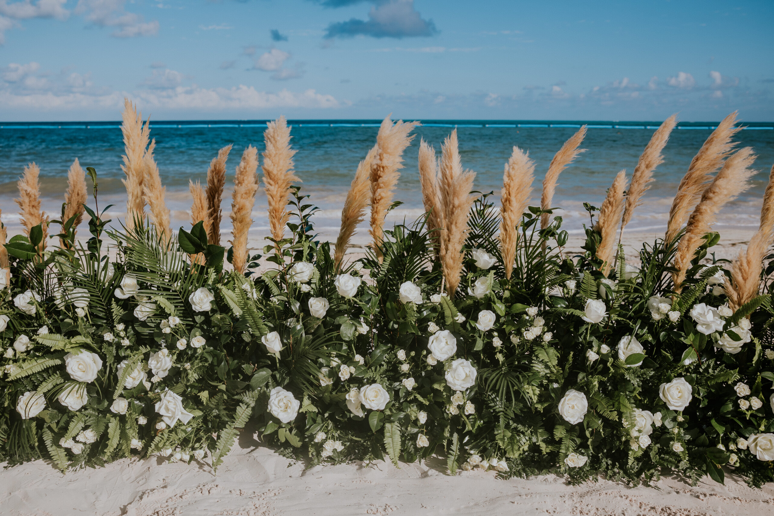 Lush floral ceremony setup with pampas grass and white roses on the sandy beach, overlooking turquoise water.