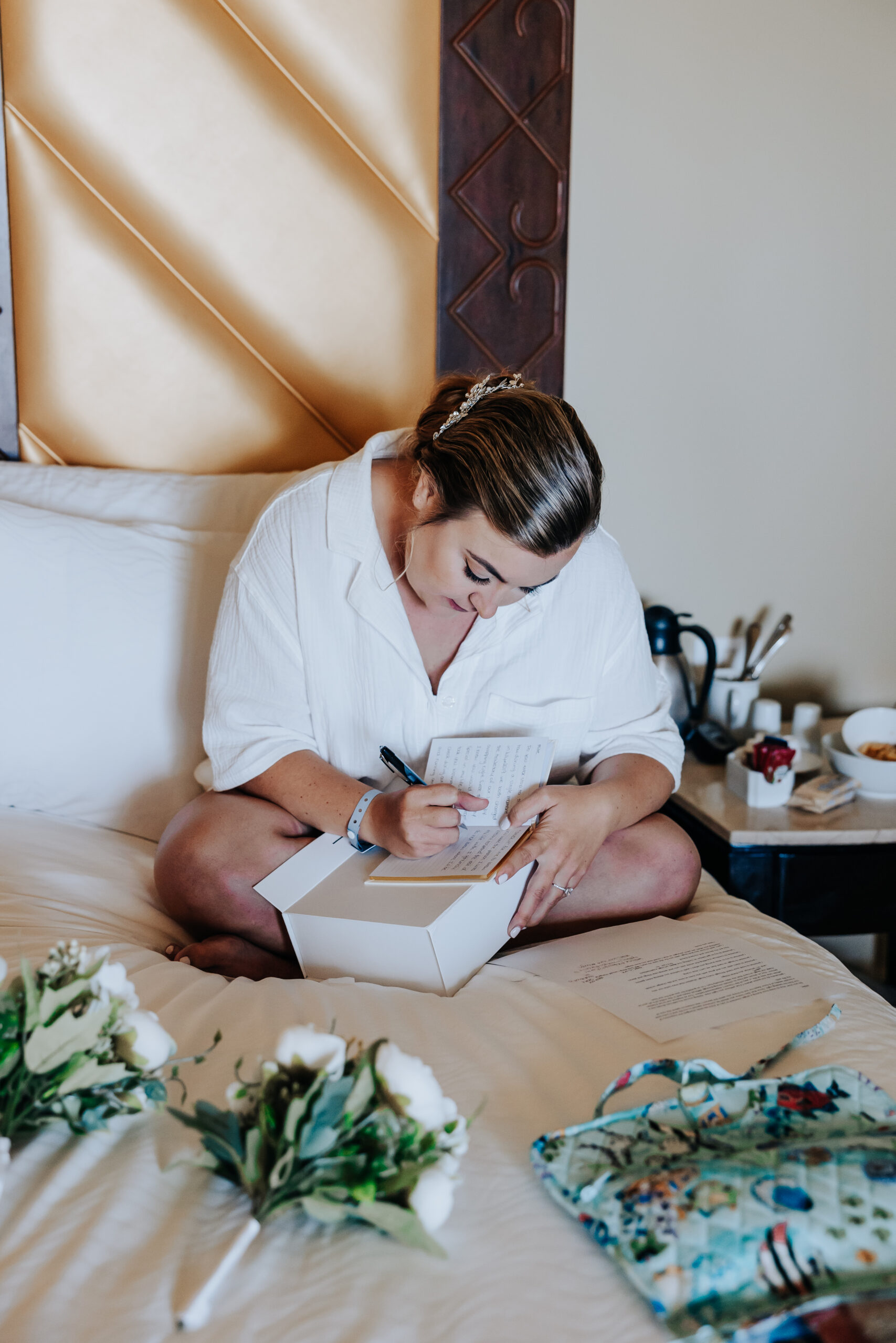 Bride sits cross-legged on the bed writing her vows, surrounded by flowers and thoughtful wedding day details.