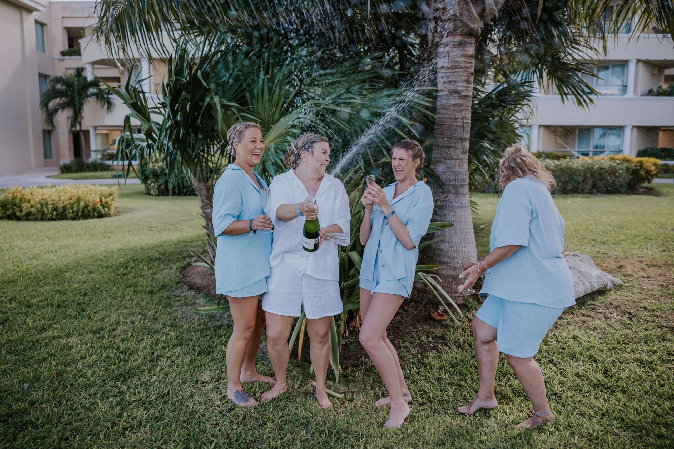 Bride pops a bottle of champagne with her girls, mid-spray, in a fun and wild celebration on the lawn.