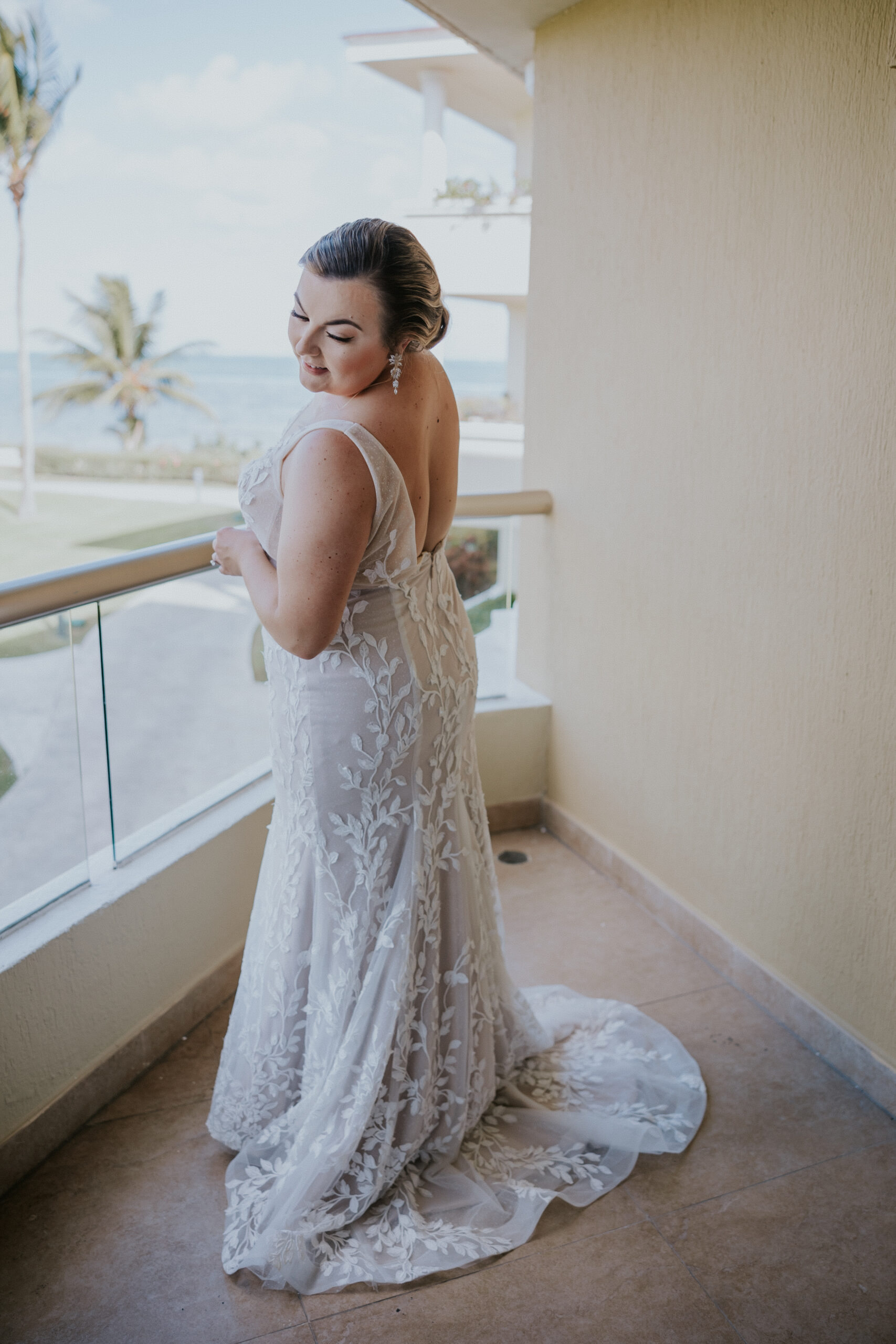Bride stands on a balcony overlooking the ocean, showing off the delicate lace detailing of her dress at a Mexico destination wedding.