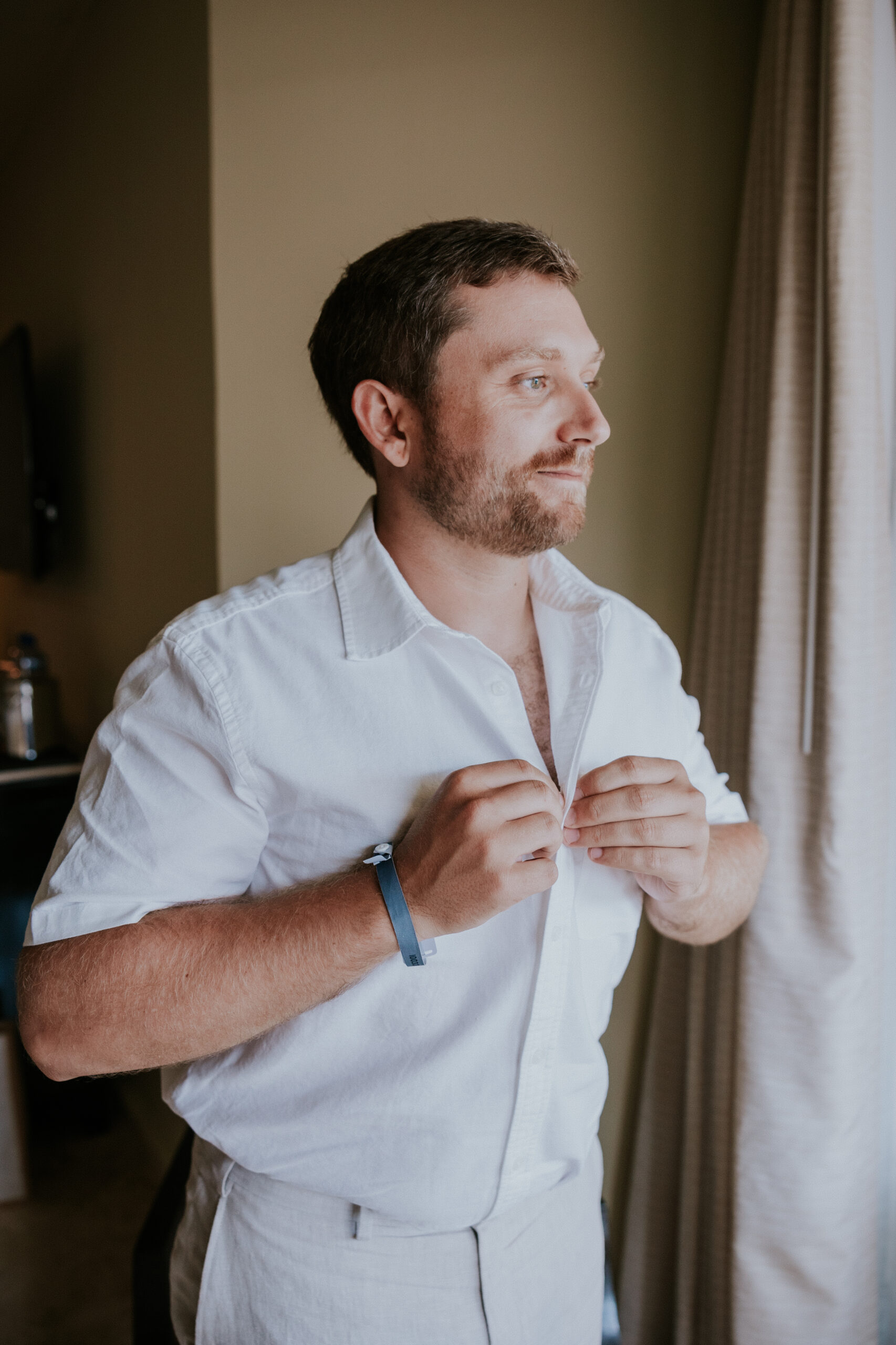Groom buttons his shirt near the window, getting ready for his Mexico destination wedding with a relaxed, tropical vibe.