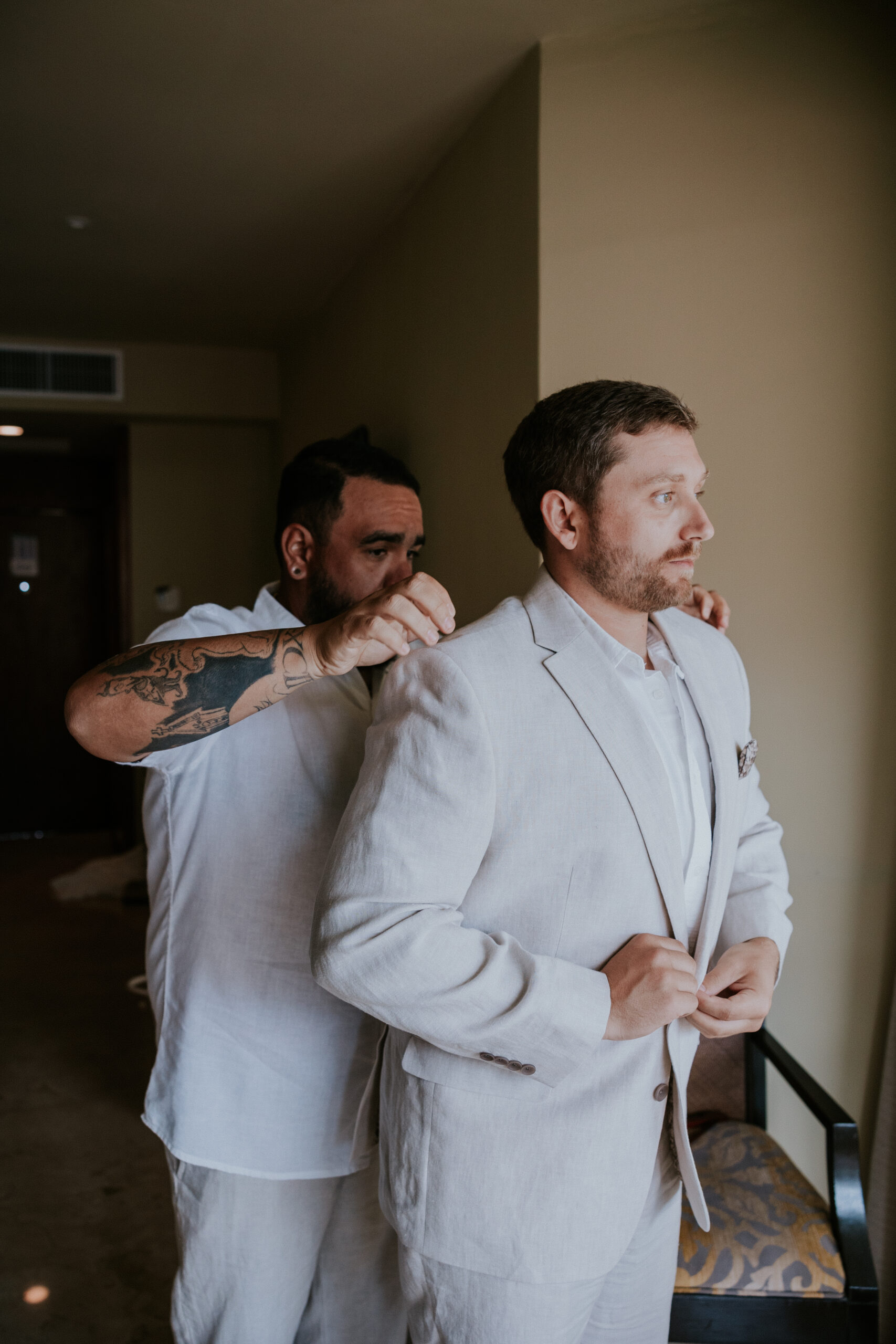 Groom’s friend helps him get ready, adjusting his collar in the hotel room before the ceremony.