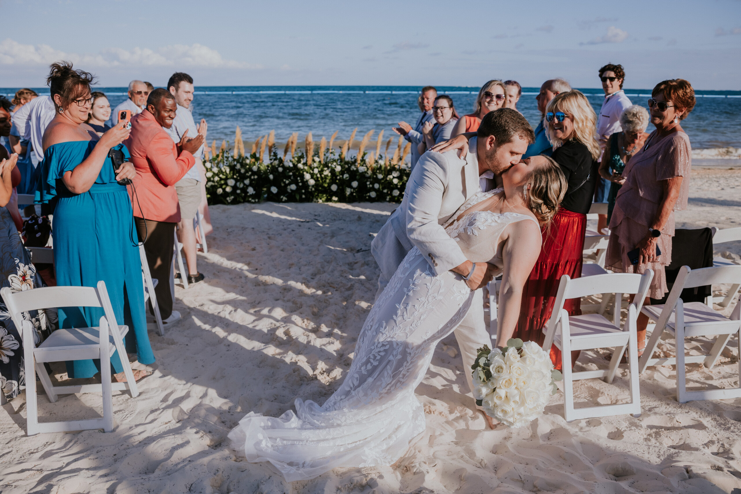 Bride and groom kiss mid-aisle as guests cheer them on during their beachside wedding celebration.