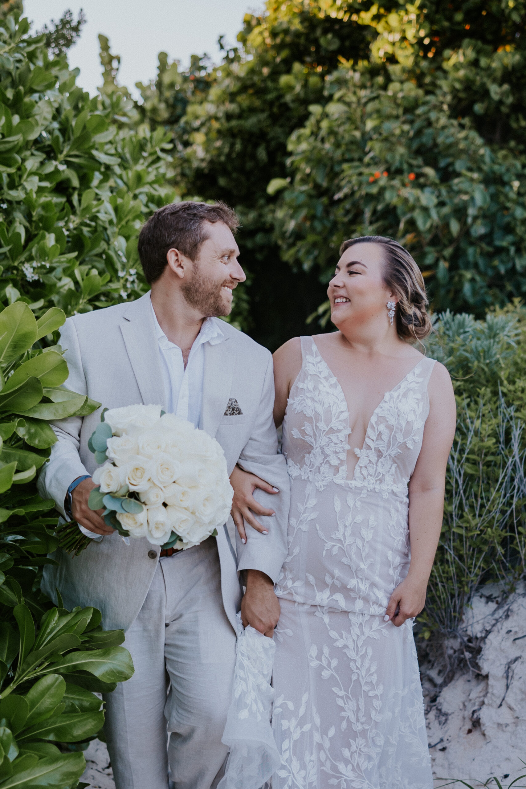 Bride and groom smiling at each other while walking through lush greenery, holding hands and a bouquet of white flowers.