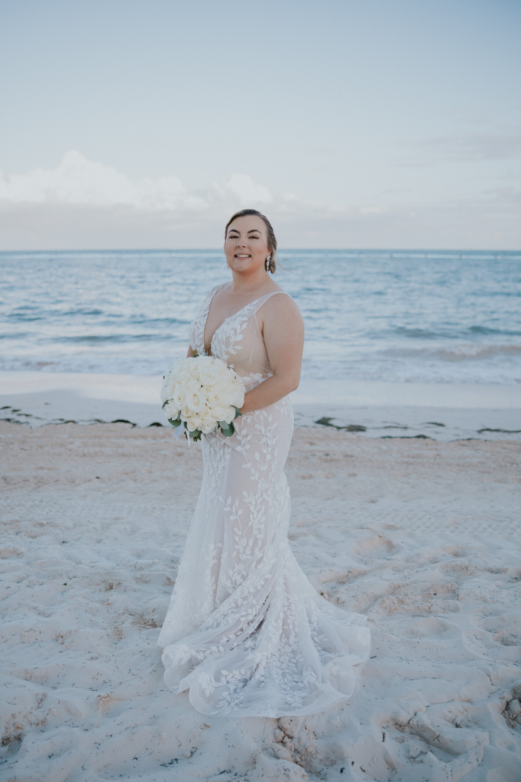 Bride in an embroidered gown holding a bouquet of white roses, standing gracefully on the beach at her Mexico destination wedding.